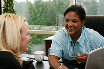 Photograph of two women smiling and talking across a desk in front of a large window.